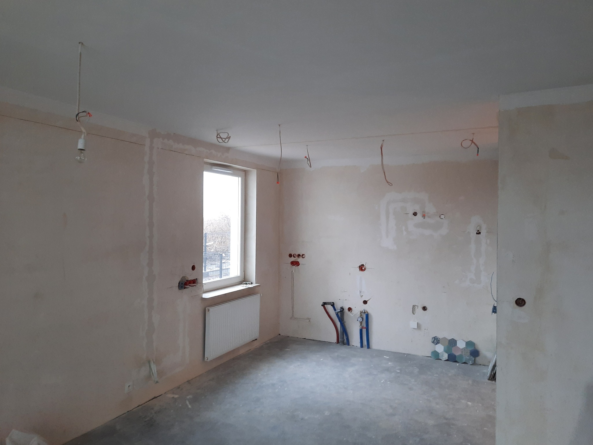 Wnętrze w trakcie remontu, widoczne przygotowane punkty elektryczne i hydrauliczne na ścianach, surowy beton na podłodze, grzejnik pod oknem, oraz wiszące kable elektryczne z żarówką.