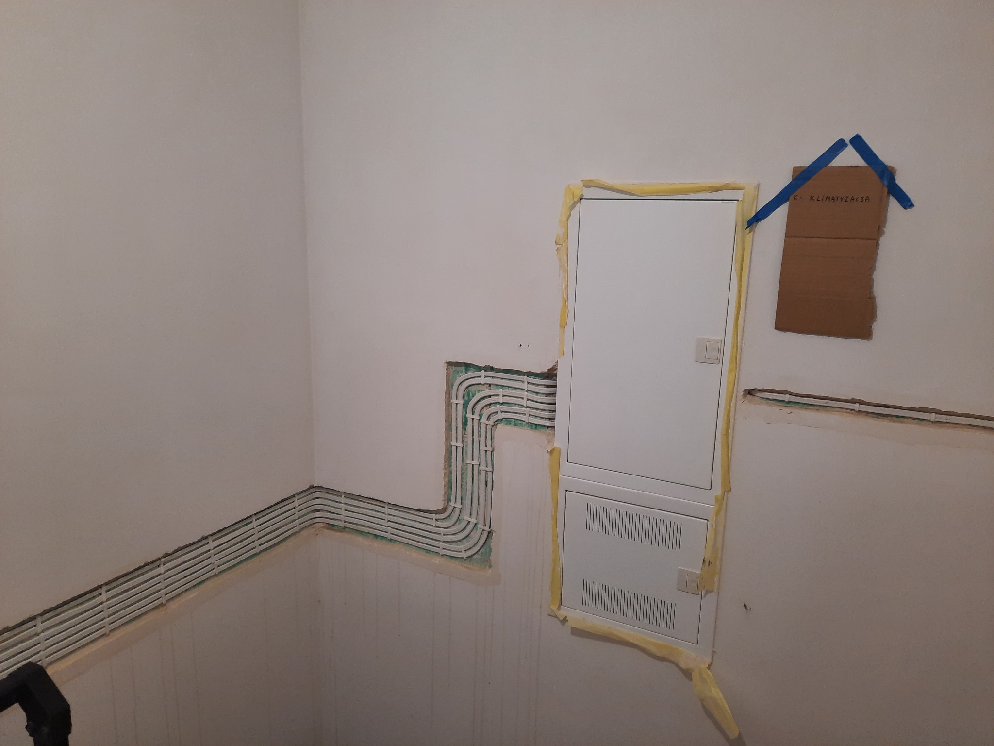Ściana w trakcie remontu z zamontowanymi białymi korytkami kablowymi i białą skrzynką elektryczną zabezpieczoną taśmą malarską, na ścianie kartka z napisem 'K-KLIMATYZACJA' przyklejona niebieską...