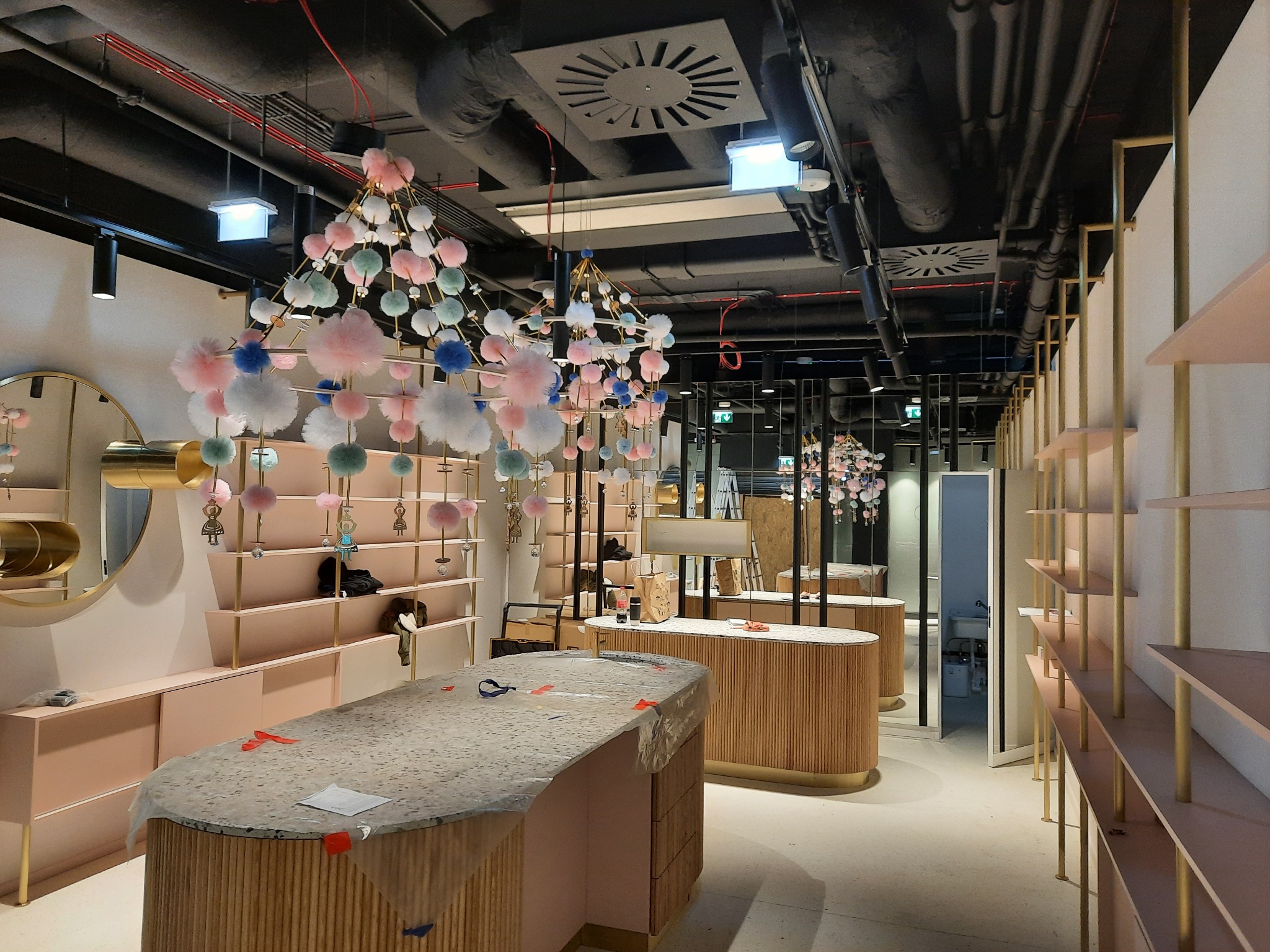 Wnętrze sklepu w trakcie wykańczania: instalacja oświetlenia dekoracyjnego z pomponami, widoczne regały, lady pod folią ochronną, sufit z instalacjami i oświetleniem technicznym.