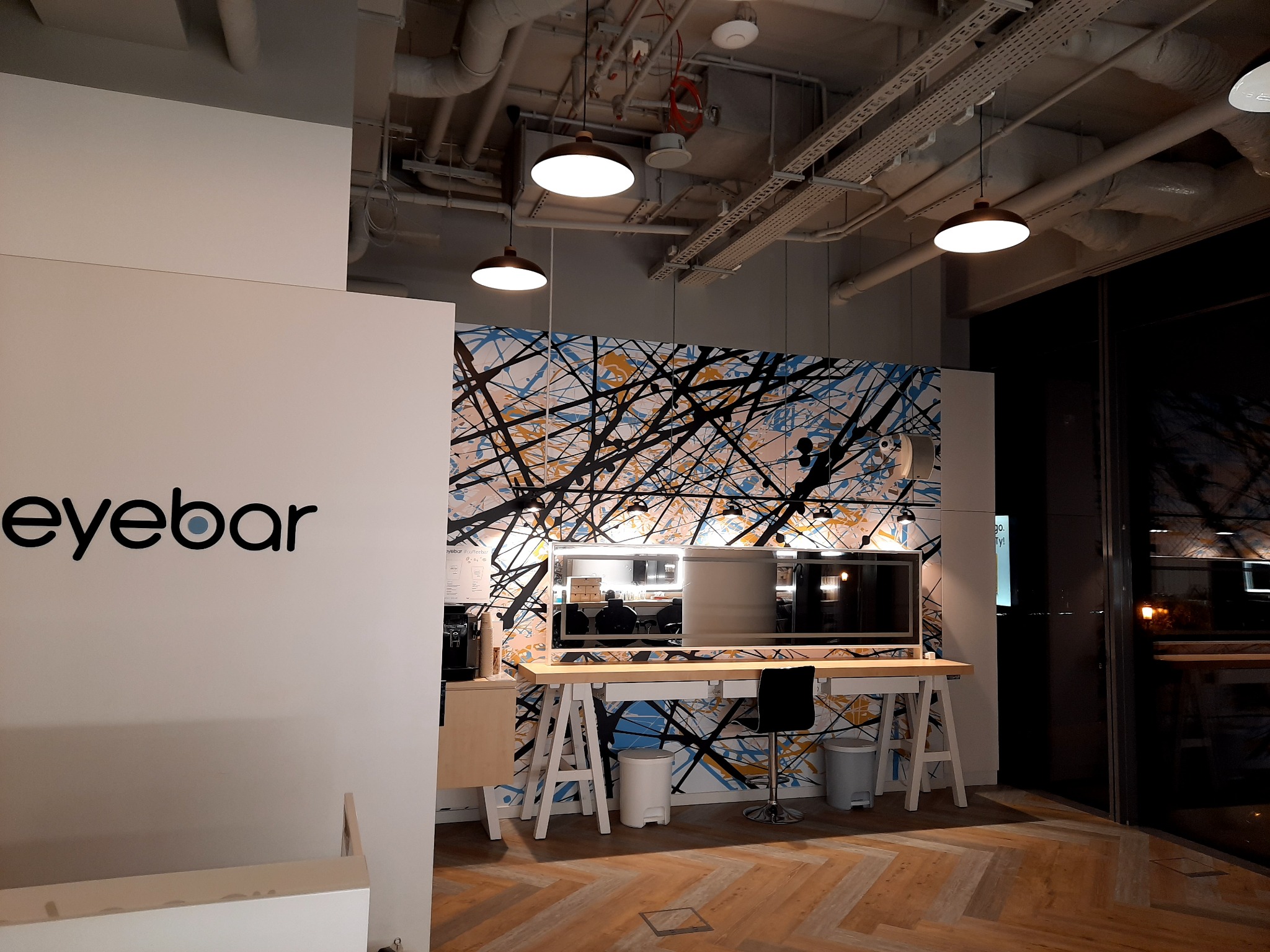 Nowoczesne biuro z logo 'eyebar' na ścianie, designerskie oświetlenie sufitowe, drewniana podłoga w jodełkę i stanowisko pracy z monitorami na tle abstrakcyjnej tapety.
