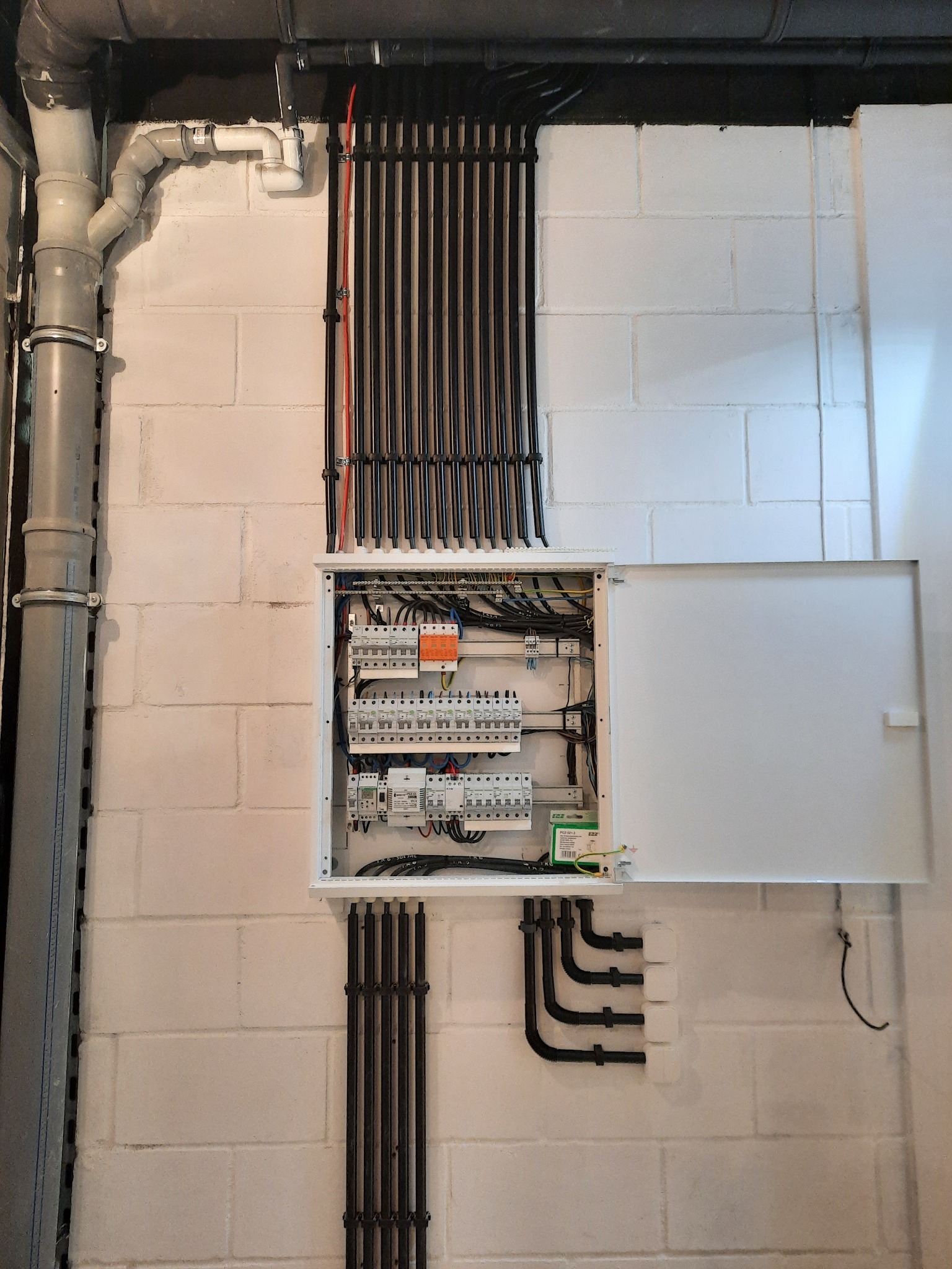 Otwarta skrzynka rozdzielcza z widocznymi bezpiecznikami i przewodami, zamontowana na pomalowanej na biało ścianie z bloczków betonowych. Rury instalacyjne w kolorze czarnym prowadzone pionowo...