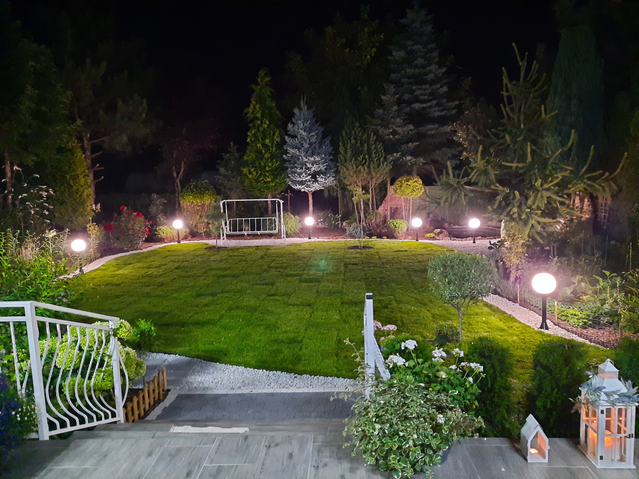 Oświetlony nocą, zadbany ogród z równo przystrzyżonym trawnikiem, ozdobnymi krzewami i lampami kulami wzdłuż ścieżki. W tle widoczna huśtawka ogrodowa.
