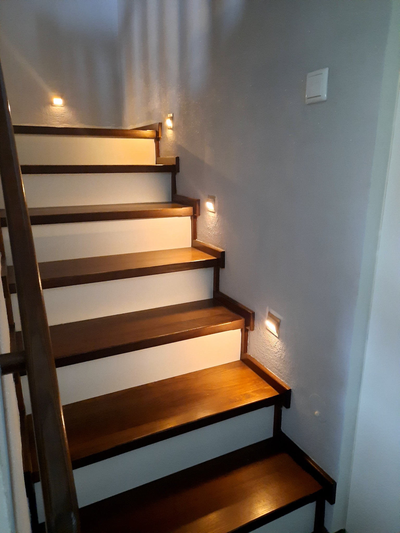 Drewniane schody z białymi podstopnicami, oświetlone małymi, kwadratowymi lampkami ściennymi, zamontowanymi przy każdym stopniu. Widoczny włącznik światła na szarej ścianie.