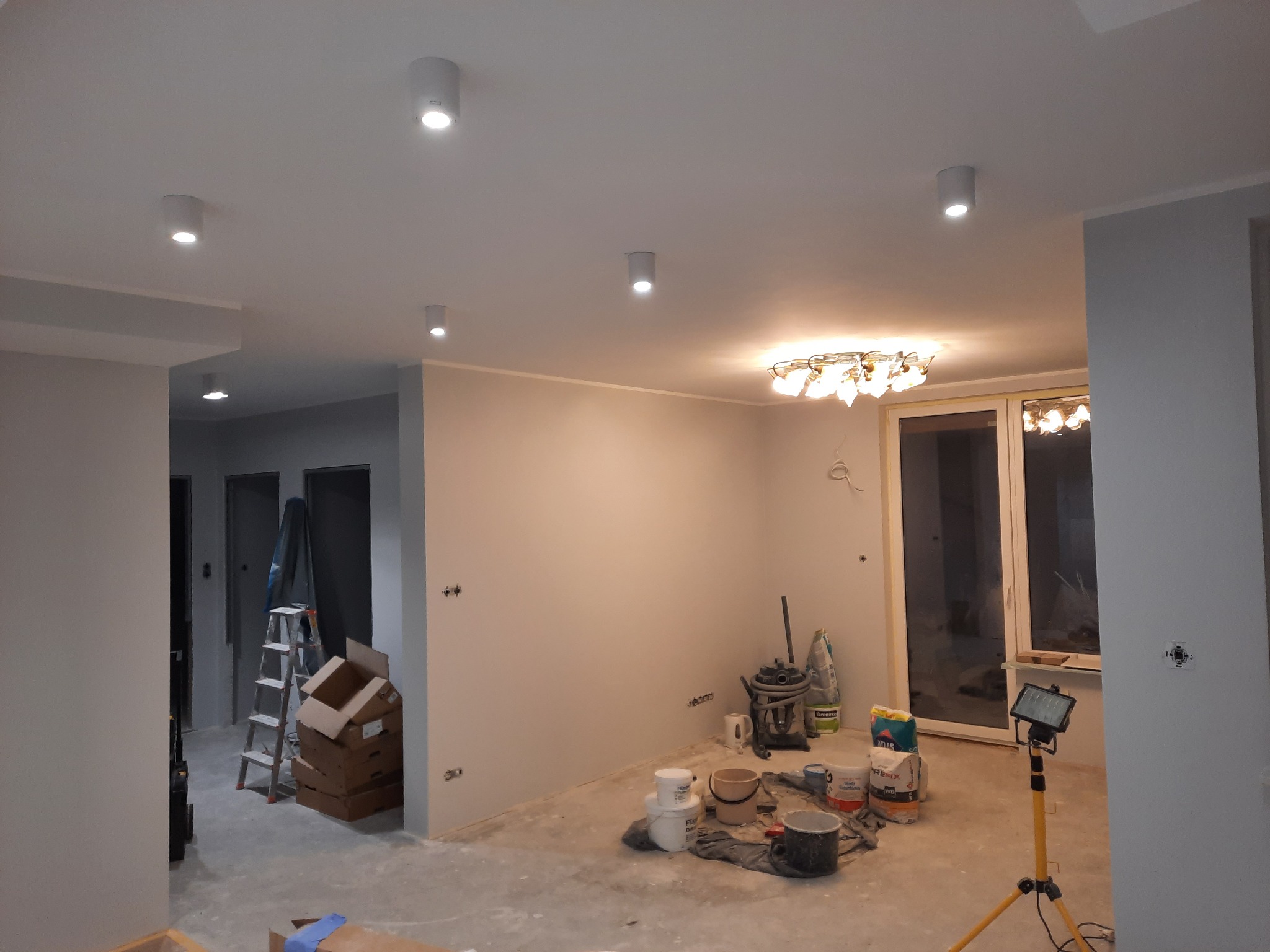Wnętrze w trakcie remontu, widoczne puszki elektryczne w ścianie, drabina, kartony, lampy sufitowe i halogen na statywie.