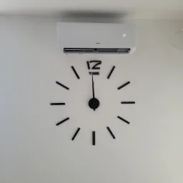 Biały klimatyzator ścienny marki AUX zamontowany nad dużym, dekoracyjnym zegarem na białej ścianie. Na wyświetlaczu klimatyzatora widoczna temperatura 32 stopnie.