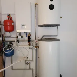 Zestaw urządzeń grzewczych: biały bojler Rotenso z dwoma czarnymi otworami inspekcyjnymi, biały panel Aquami i czerwony zbiornik wyrównawczy, połączone rurami z zaworami i manometrami.