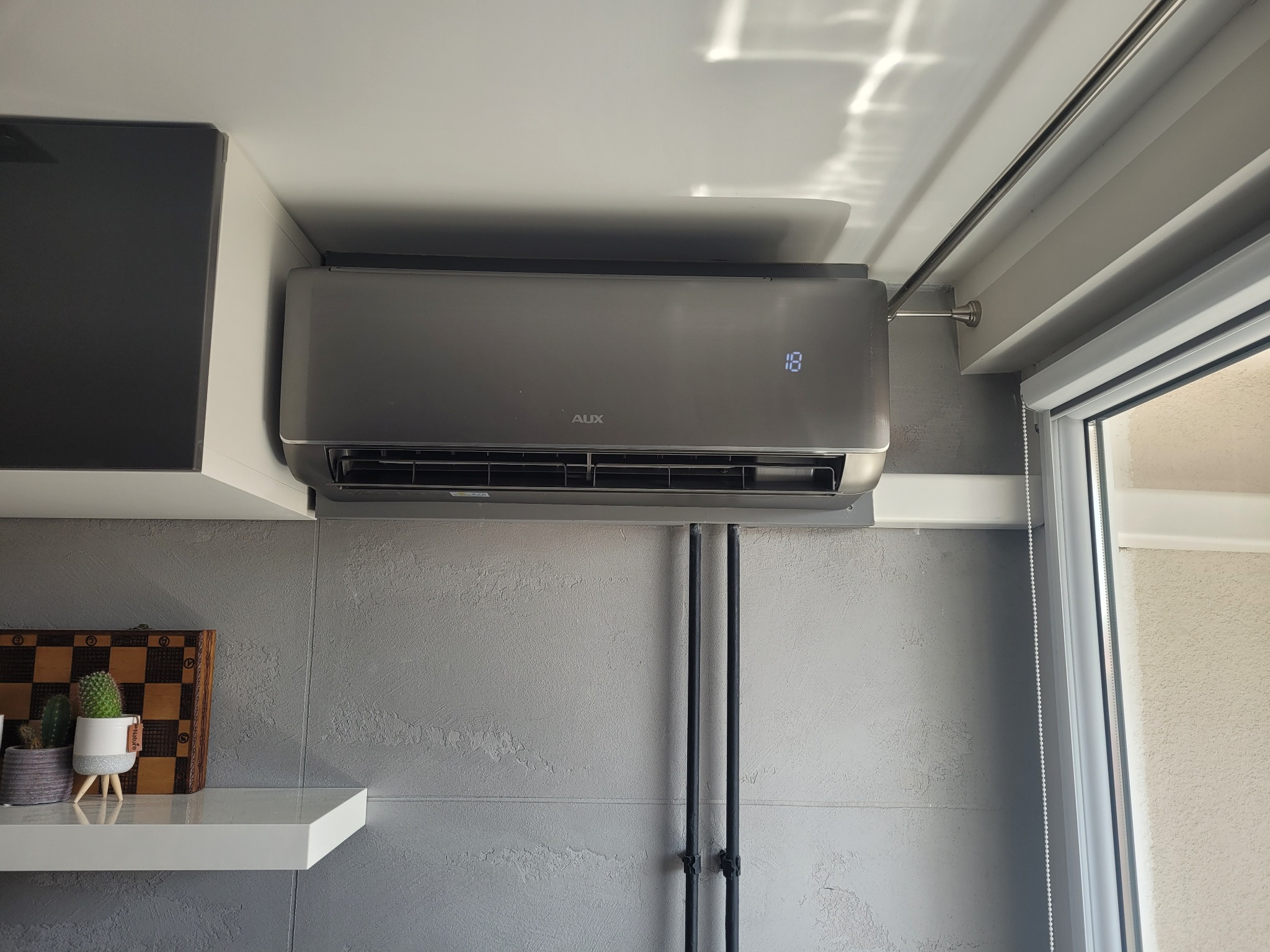 Szary klimatyzator AUX zamontowany w nowoczesnym wnętrzu, widoczny wyświetlacz z temperaturą 18 stopni, obok okno z roletami oraz półka z szachami i kaktusami.