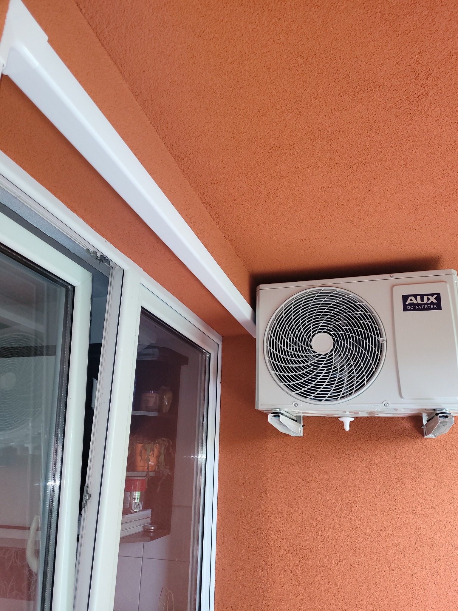 Zamontowana jednostka zewnętrzna klimatyzacji AUX DC Inverter na pomarańczowej ścianie budynku, widoczna obok białego okna z widokiem na półkę z doniczkami.