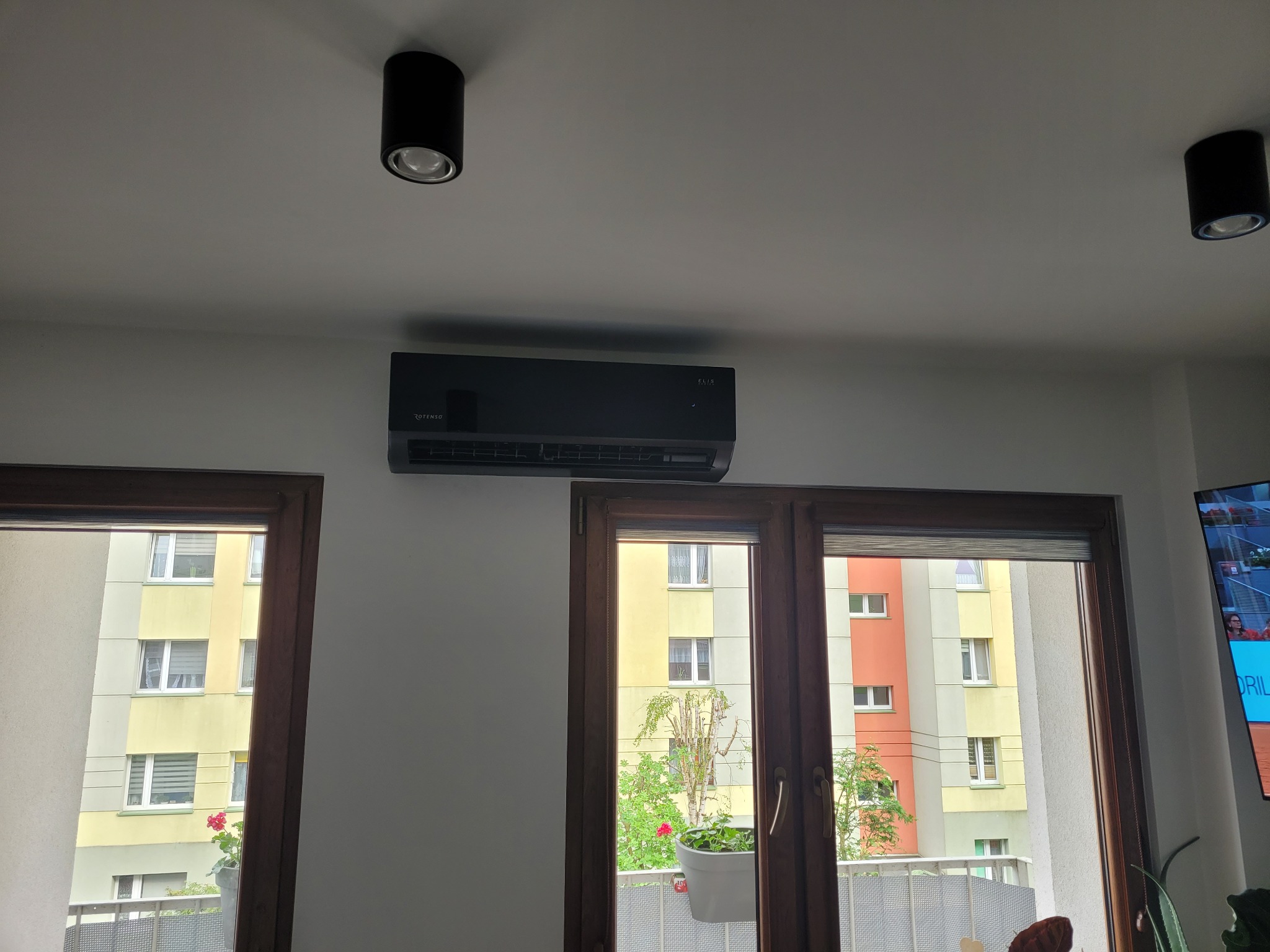 Czarny klimatyzator ścienny zamontowany nad dwoma oknami w mieszkaniu z widokiem na blok z balkonami.