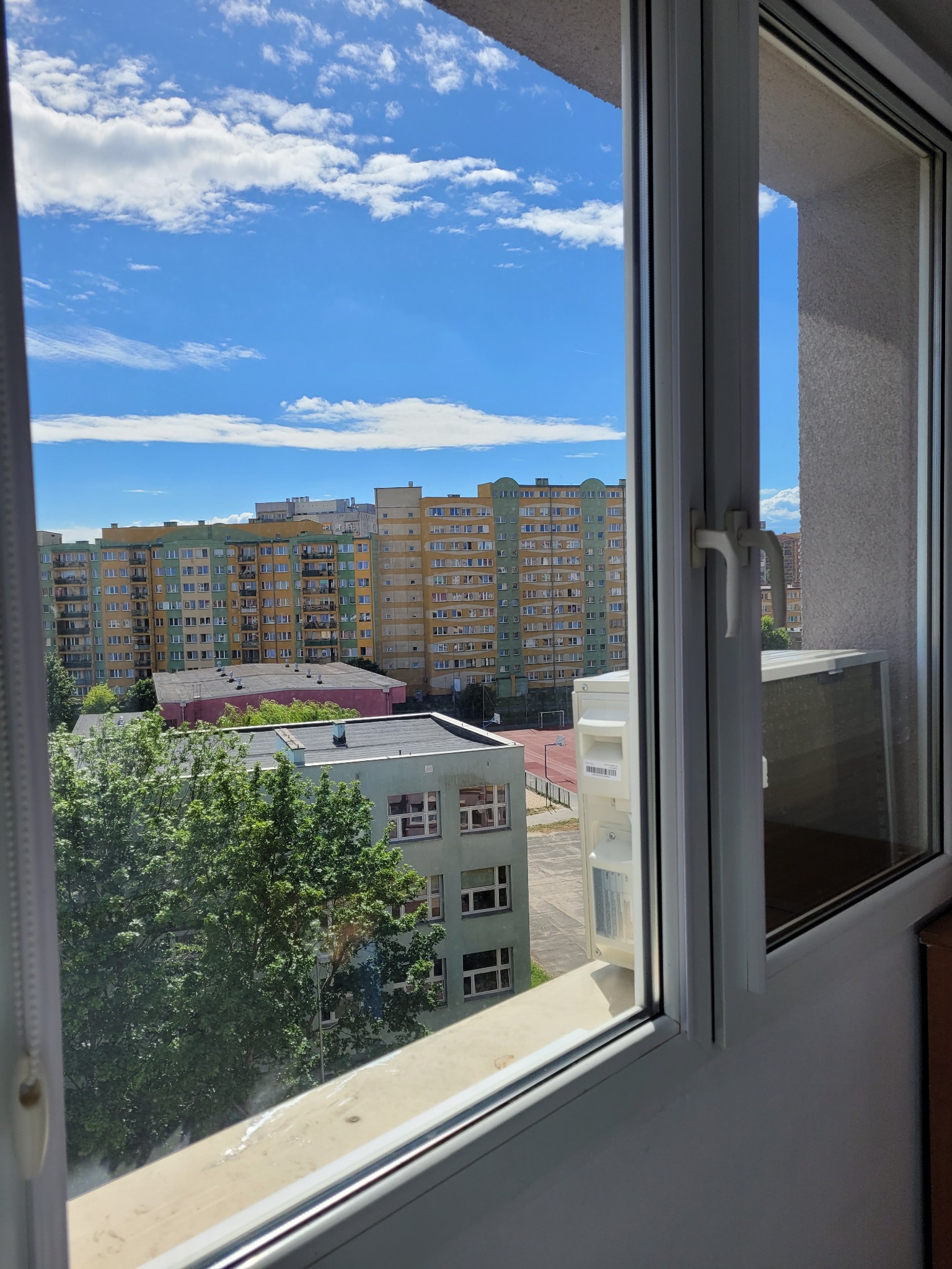 Widok z okna na osiedle z zamontowanym klimatyzatorem zewnętrznym, błękitne niebo z chmurami w tle.