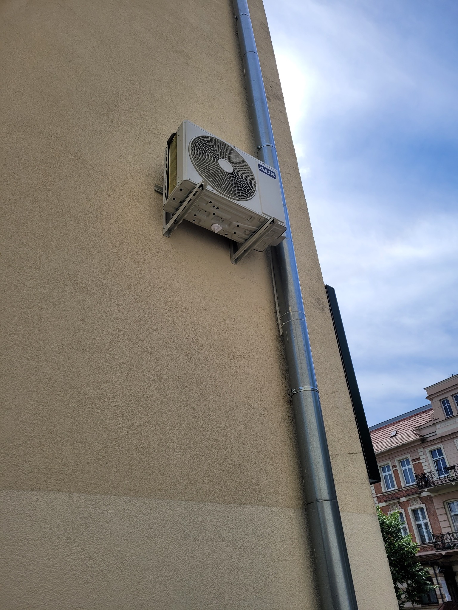 Zamontowana jednostka zewnętrzna klimatyzacji AUX na elewacji budynku, obok rury spustowej; widoczne mocowania i fragment budynku w tle.