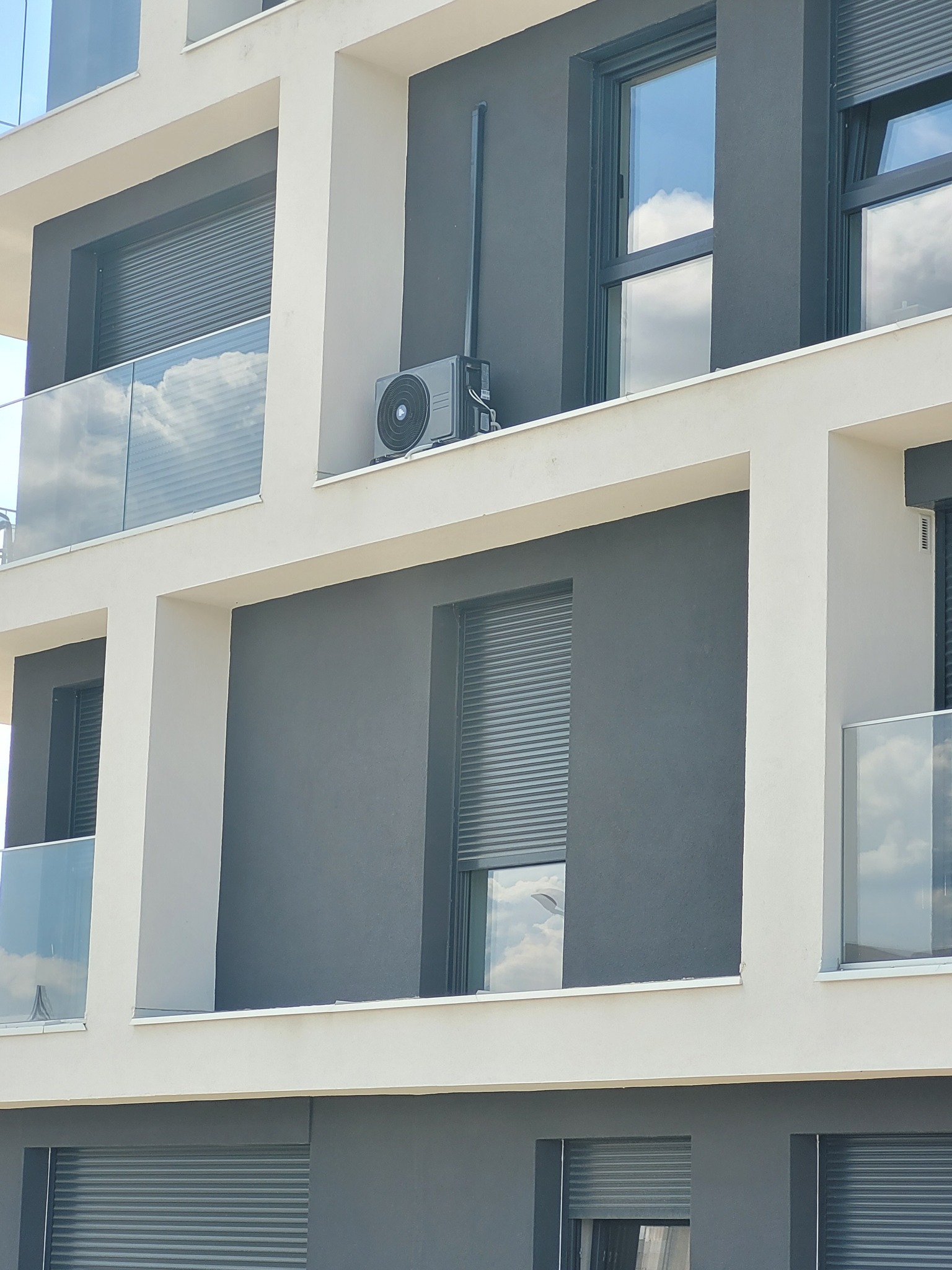 Zewnętrzna jednostka klimatyzacji zamontowana na balkonie nowoczesnego budynku mieszkalnego z szarymi ścianami i białymi elementami architektonicznymi.