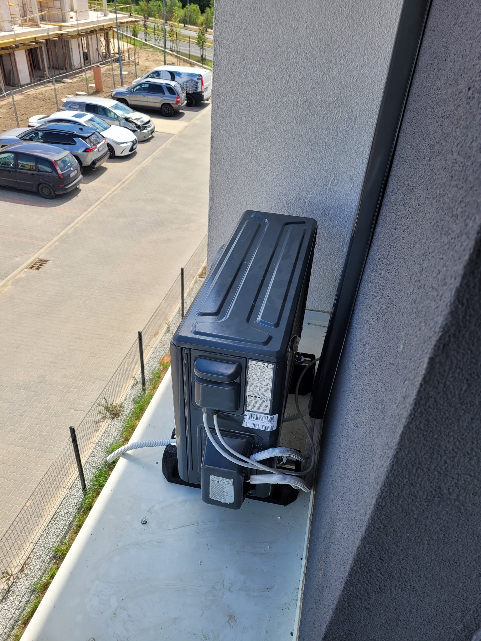 Zewnętrzna jednostka klimatyzacji Kaisai zamontowana na balkonie nowego budynku mieszkalnego, widok z góry na osiedle i parking.