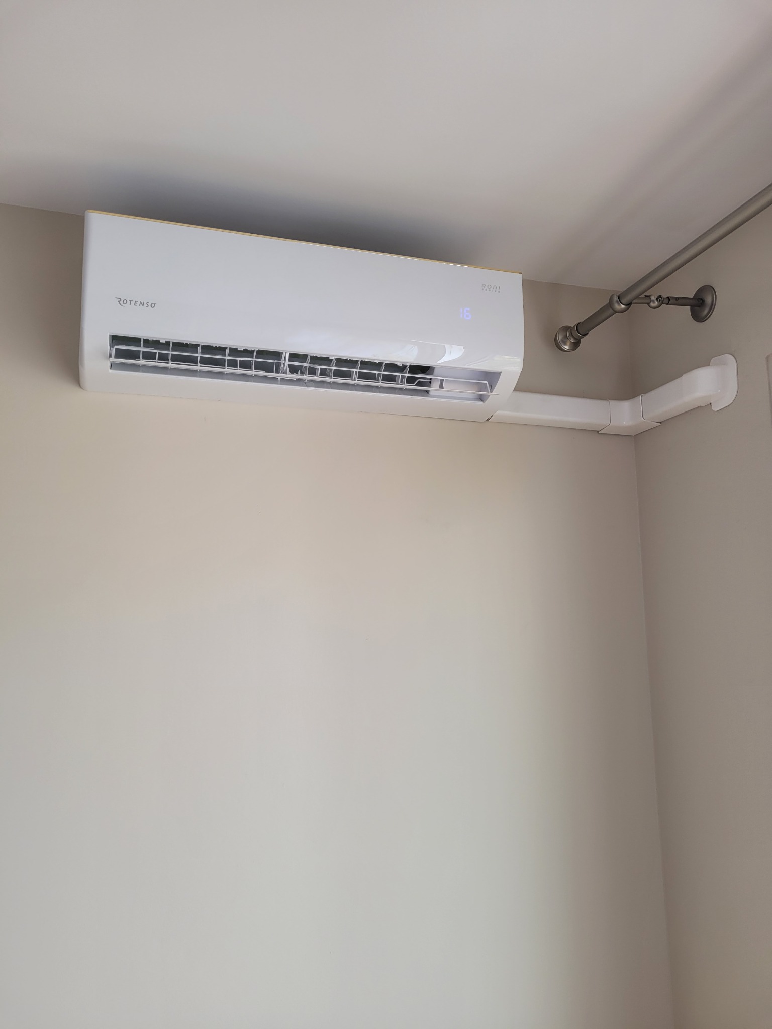 Biały klimatyzator Rotenso zamontowany na beżowej ścianie w pomieszczeniu, z widoczną białą rurą odprowadzającą i fragmentem karnisza na ścianie.