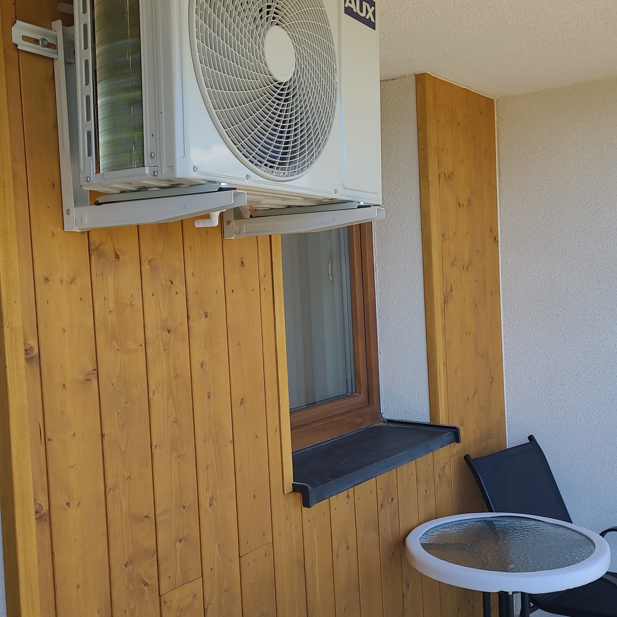 Biała jednostka zewnętrzna klimatyzacji AUX zamontowana na drewnianej elewacji balkonu, widoczny stolik i krzesło.