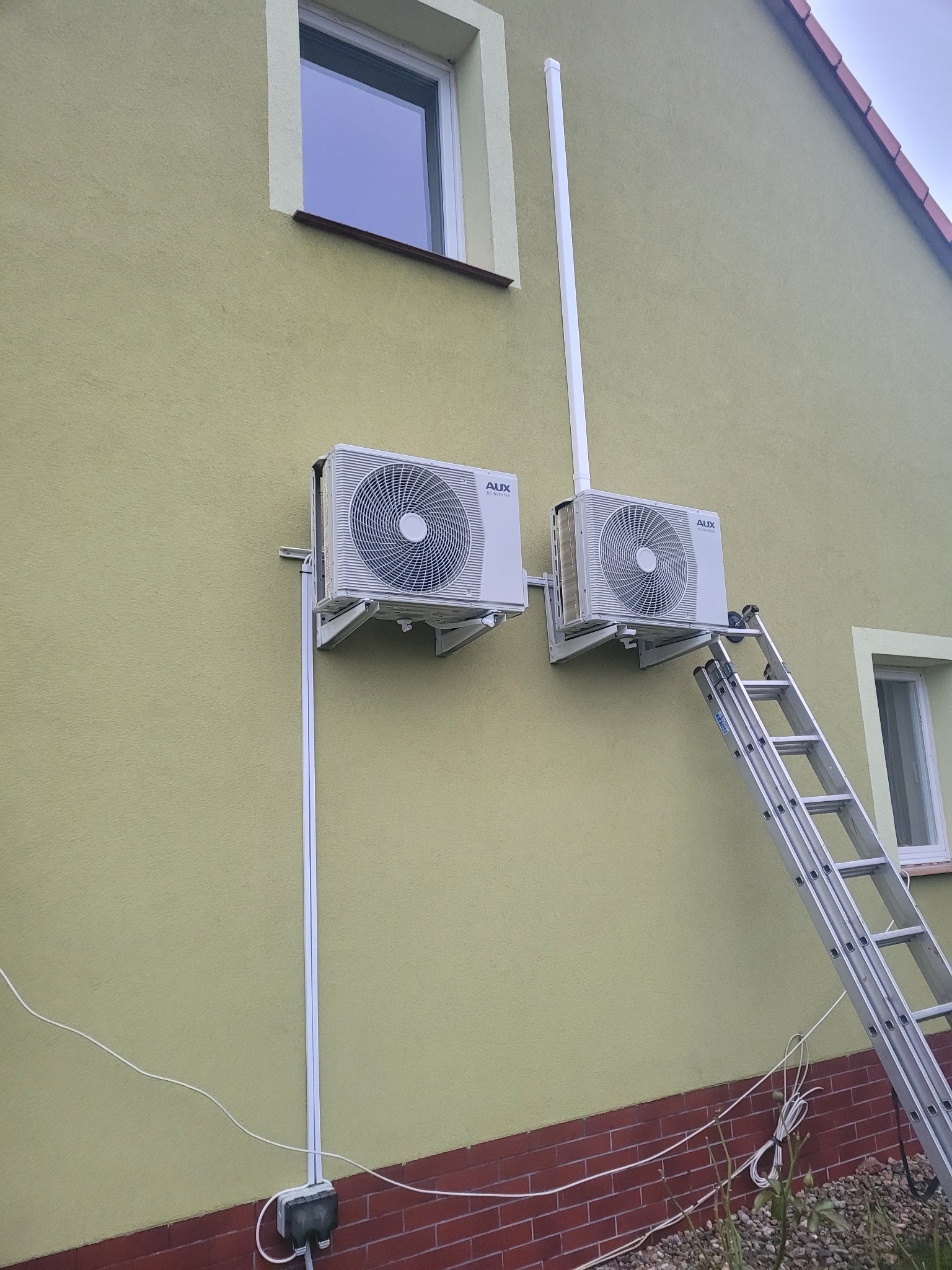 Dwa klimatyzatory marki AUX zamontowane na elewacji budynku jednorodzinnego. Do jednego z urządzeń przystawiona aluminiowa drabina. Widoczne przewody i peszle instalacyjne.