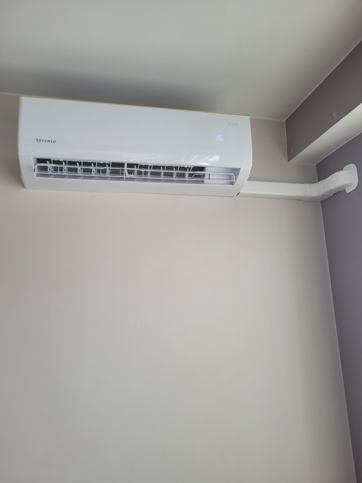 Zamontowany na ścianie biały klimatyzator Rotenso z wyświetlaczem ustawionym na 16 stopni Celsjusza, z widoczną instalacją odprowadzającą skropliny w maskownicy.