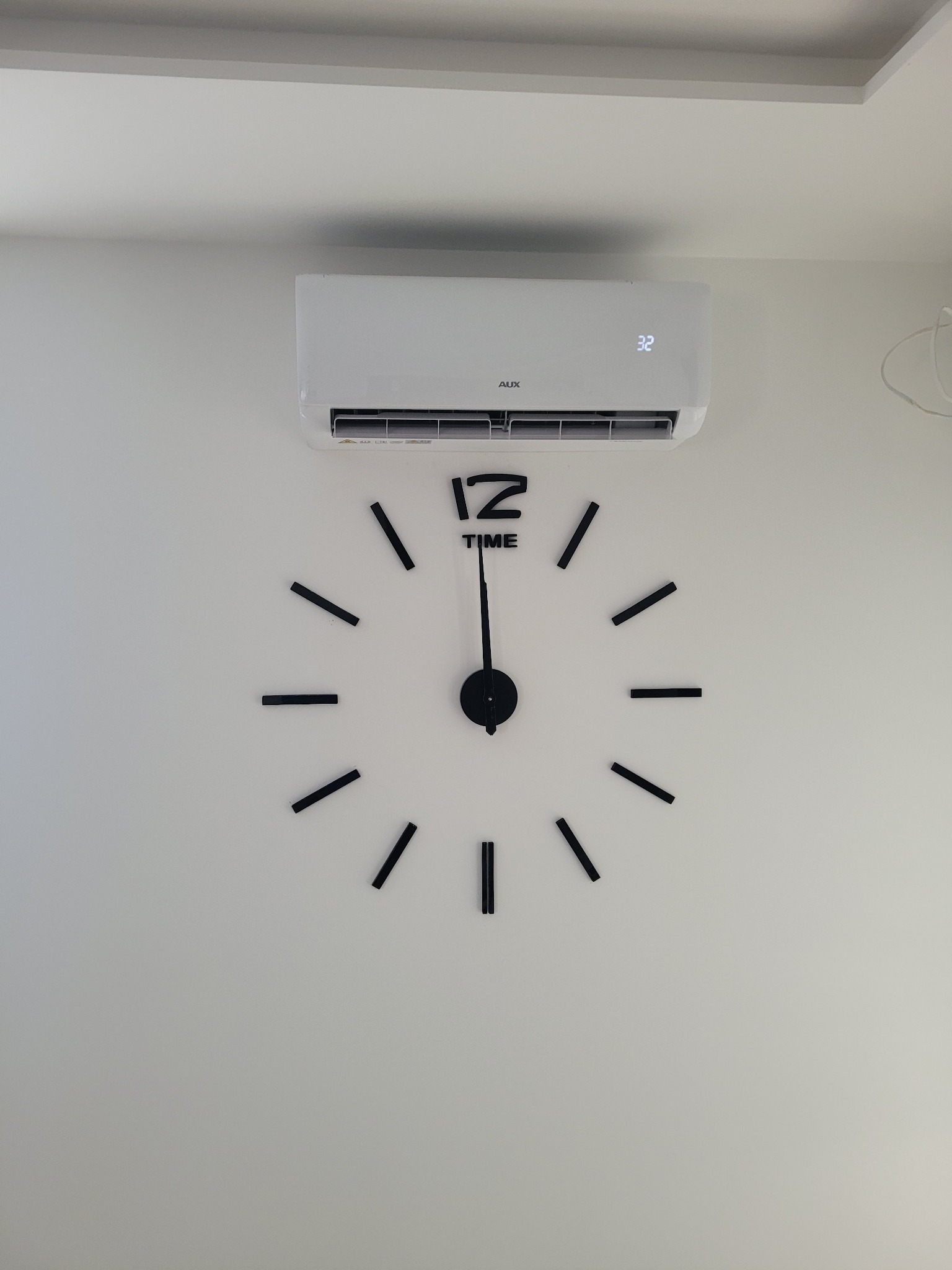 Biały klimatyzator ścienny marki AUX zamontowany nad dużym, dekoracyjnym zegarem na białej ścianie. Na wyświetlaczu klimatyzatora widoczna temperatura 32 stopnie.