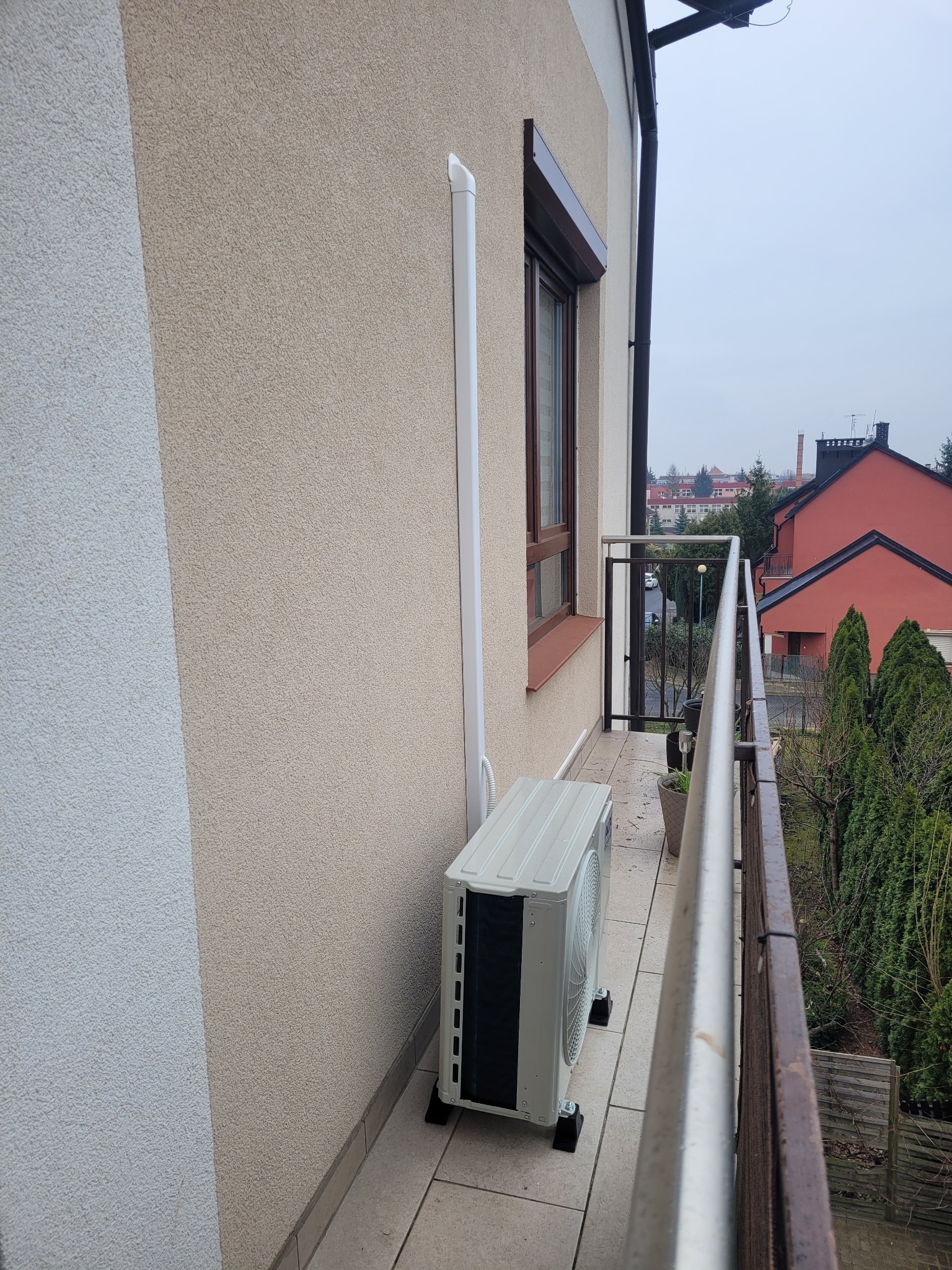 Klimatyzator zewnętrzny zamontowany na balkonie bloku mieszkalnego, z widoczną rurą odprowadzającą na elewacji i fragmentem balustrady.