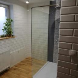 Polbud Paweł Gliszczyński - Wykończona łazienka w Inowrocławiu z kabiną prysznicową typu walk-in, jasnymi płytkami imitującymi cegłę na ścianach, drewnianą podłogą i grzejnikiem pod oknem z rośliną doniczkową.