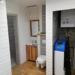 Nowoczesna łazienka z drewnianą podłogą, białymi płytkami imitującymi cegłę, szklaną kabiną prysznicową, drewnianą szafką pod umywalką, wiszącą toaletą z wzorzystą ścianką i zmiękczaczem wody obok...