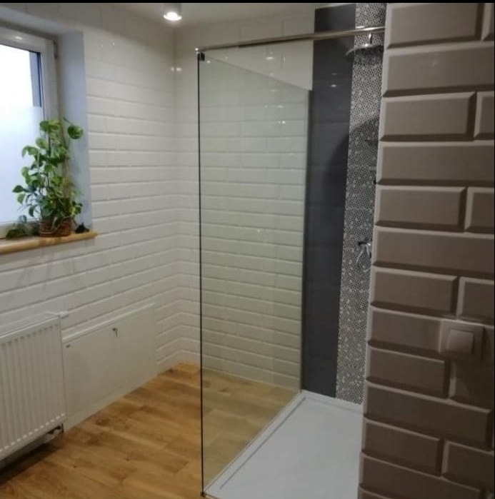Wykończona łazienka w Inowrocławiu z kabiną prysznicową typu walk-in, jasnymi płytkami imitującymi cegłę na ścianach, drewnianą podłogą i grzejnikiem pod oknem z rośliną doniczkową.