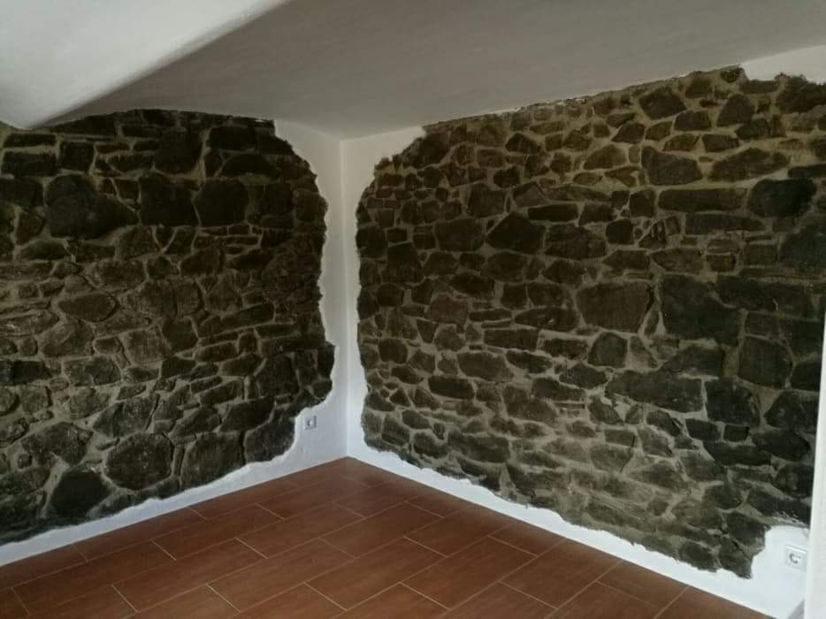 Surowe kamienne ściany w pomieszczeniu z brązową podłogą imitującą drewno; widoczne białe fragmenty ściany i sufitu.