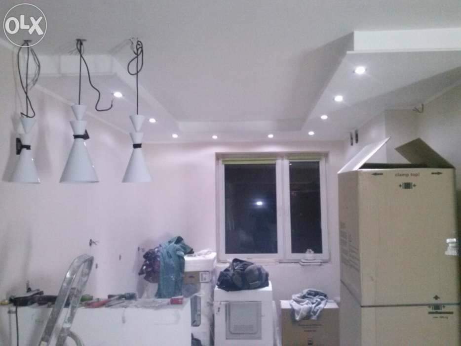 Wnętrze pokoju w trakcie remontu z trzema nowoczesnymi białymi lampami wiszącymi na tle sufitu podwieszanego z oświetleniem punktowym, widoczne narzędzia i materiały budowlane oraz kartony.