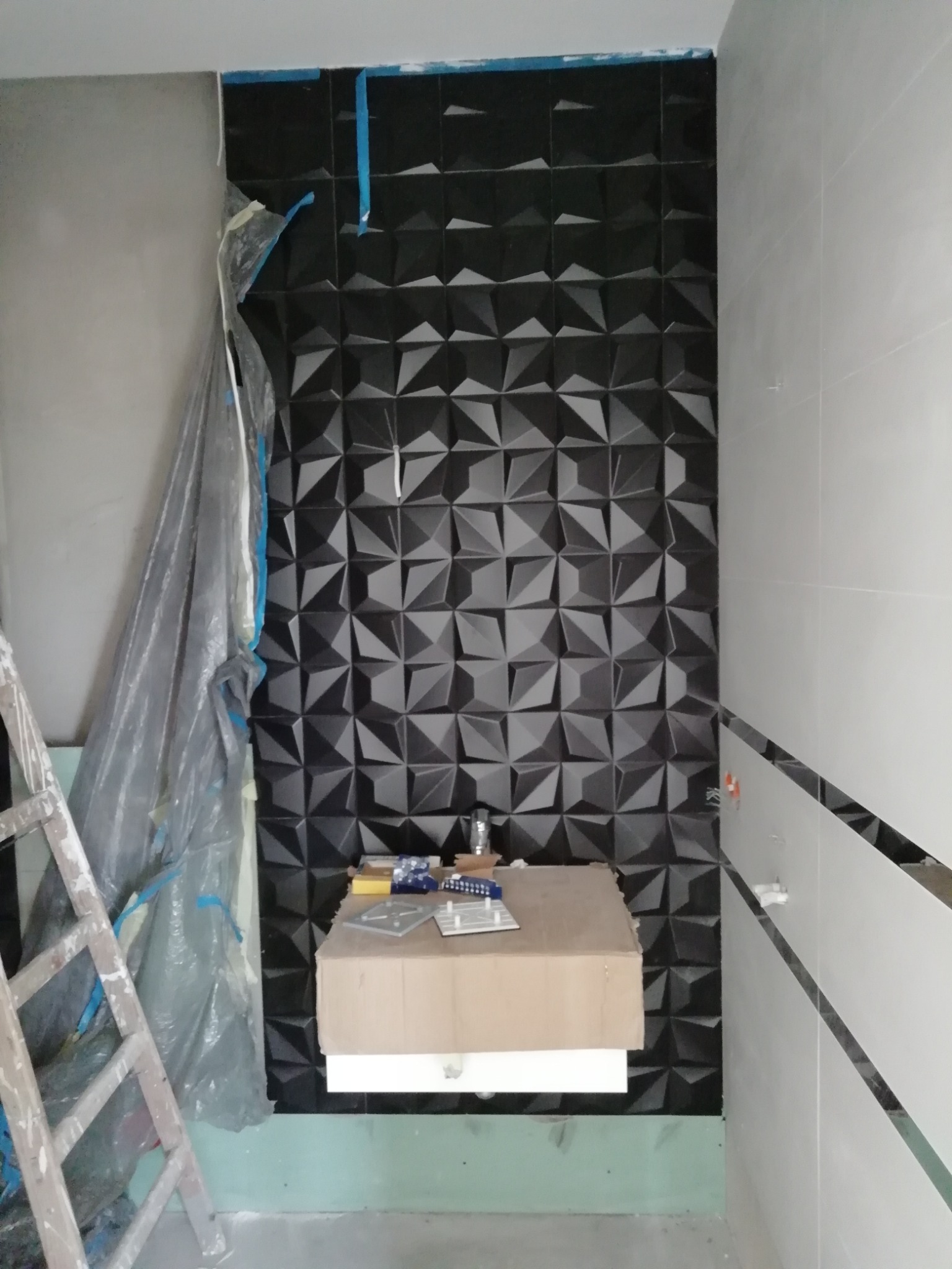 Montaż dekoracyjnej, trójwymiarowej, czarnej okładziny ściennej w łazience, z częściowo zabezpieczoną ścianą folią malarską i rozstawioną drabiną.