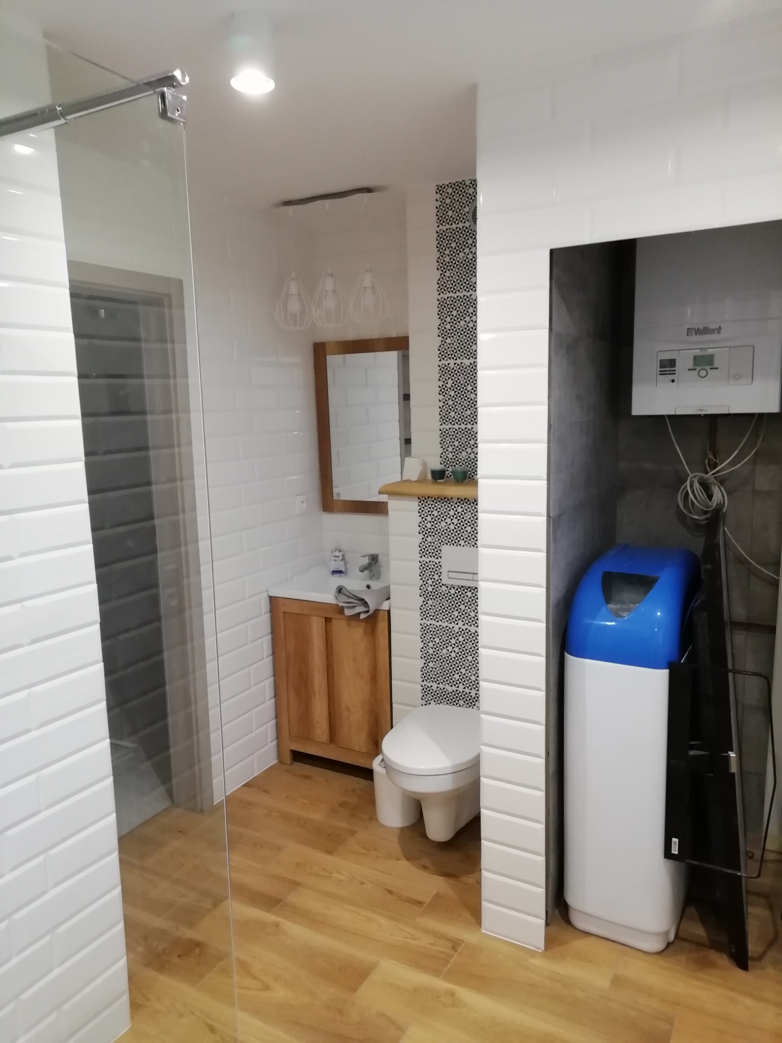 Nowoczesna łazienka z drewnianą podłogą, białymi płytkami imitującymi cegłę, szklaną kabiną prysznicową, drewnianą szafką pod umywalką, wiszącą toaletą z wzorzystą ścianką i zmiękczaczem wody obok...