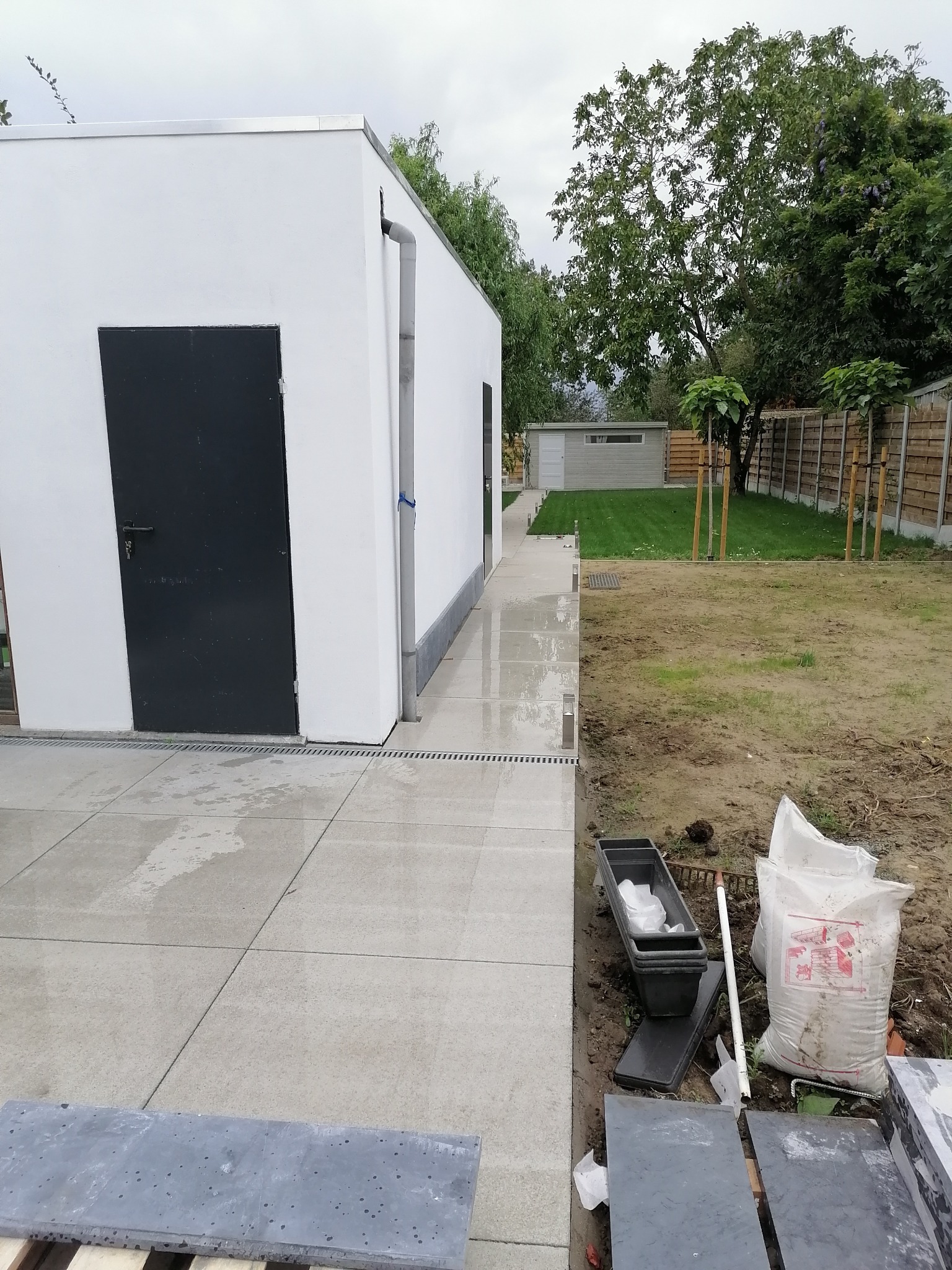 Biały budynek z ciemnymi drzwiami i nową szarą kostką brukową, widoczne materiały budowlane i narzędzia ogrodnicze na ziemi.