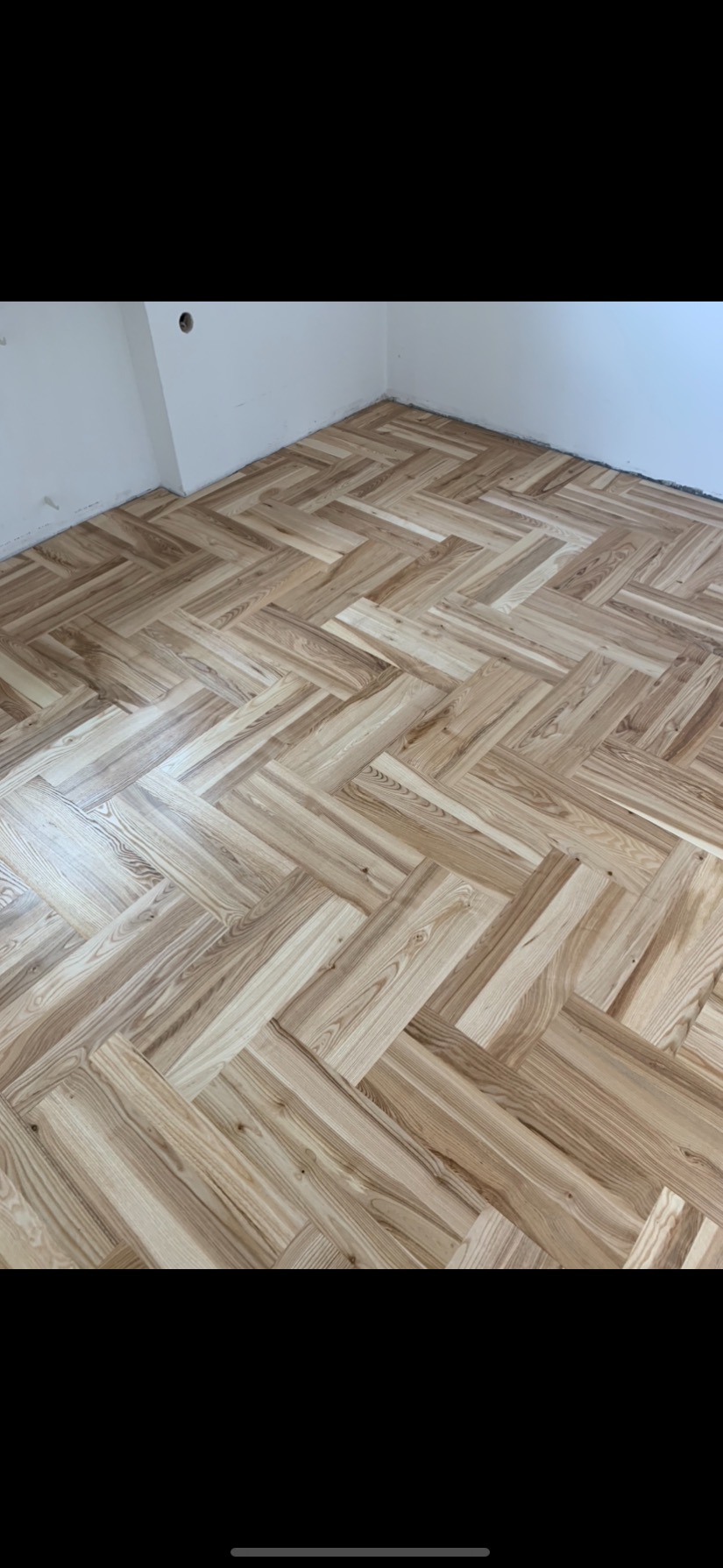 Jasny parkiet ułożony we wzór jodełki francuskiej, widoczne niedoskonałości przy ścianie, białe ściany z widocznym gniazdkiem elektrycznym.