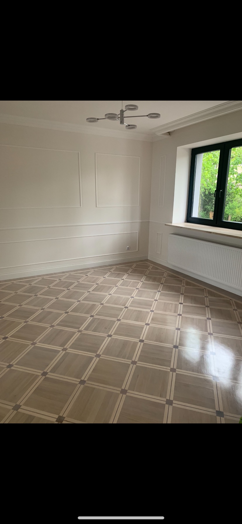 Wnętrze pokoju z nową, błyszczącą podłogą ułożoną w kwadratowy wzór, jasne ściany z dekoracyjnymi listwami, nowoczesna lampa sufitowa i okno z widokiem na zieleń.