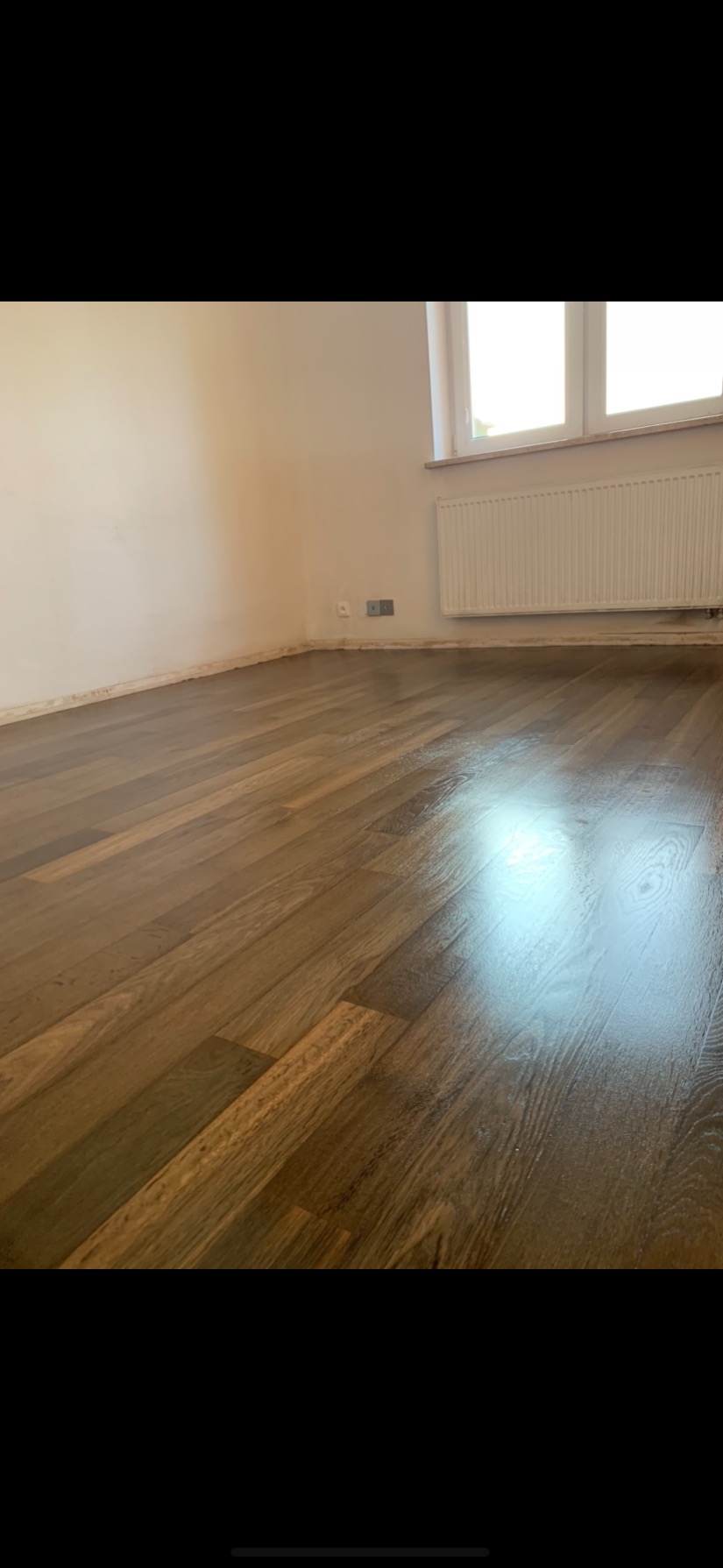 Świeżo położony, lakierowany parkiet z widocznymi śladami po pracach wykończeniowych przy ścianach, jasne wnętrze z grzejnikiem pod oknem.