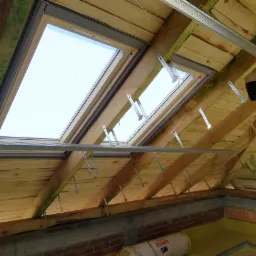 Widok od wewnątrz na drewnianą konstrukcję dachu z zamontowanymi oknami dachowymi, widoczne metalowe wsporniki, częściowo ocieplony strop i fragment ściany z cegły.