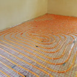 Pomarańczowe rurki ogrzewania podłogowego ułożone w pętle na siatce z folią odblaskową w rogu pomieszczenia, zabezpieczone białymi uchwytami.