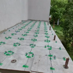 Układanie jasnych płytek na tarasie z użyciem zielonych klipsów poziomujących, widoczne otwory na słupki i fragment nieukończonej krawędzi.