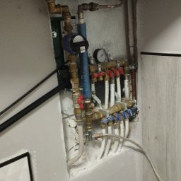 Glazurnik - Węzeł wodny z licznikiem, manometrem i zaworami w szarej wnęce. Widoczne filtry wody, rury miedziane i plastikowe. Instalacja połączona z siecią wodociągową.