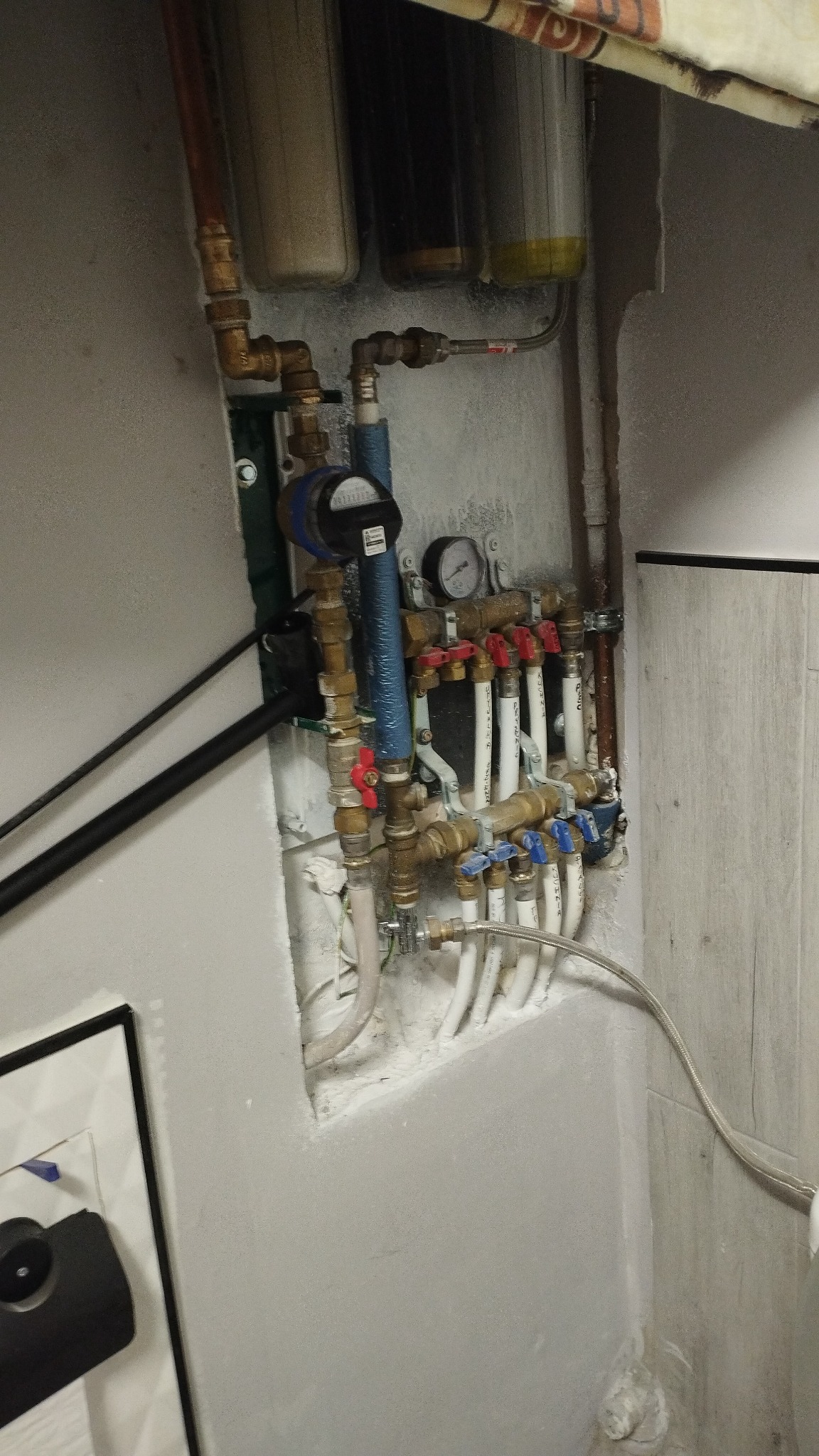 Węzeł wodny z licznikiem, manometrem i zaworami w szarej wnęce. Widoczne filtry wody, rury miedziane i plastikowe. Instalacja połączona z siecią wodociągową.