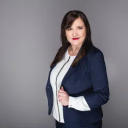 Portret kobiety w granatowej marynarce i białej bluzce z ozdobnym czarnym paskiem, na jednolitym szarym tle, z delikatnym uśmiechem i ciemnymi włosami opadającymi na ramiona.