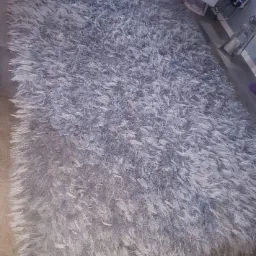 Jasnoszary, puszysty dywan typu shaggy na drewnianej podłodze, widok z góry pod kątem, z elementami wyposażenia wnętrza w tle.