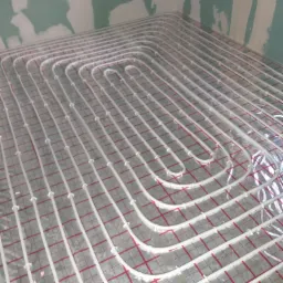 Białe rury ogrzewania podłogowego ułożone na folii z czerwoną kratką, widok z góry, ściany w tle w stanie surowym.