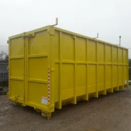 Żółty kontener na gruz z widocznymi kółkami transportowymi, ustawiony na żwirowym podłożu, w tle siatka ogrodzeniowa i zarośla.