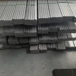 Stosy nierdzewnych, płaskich profili, zabezpieczonych zielonymi taśmami, ułożone na szarym, betonowym podłożu w magazynie.