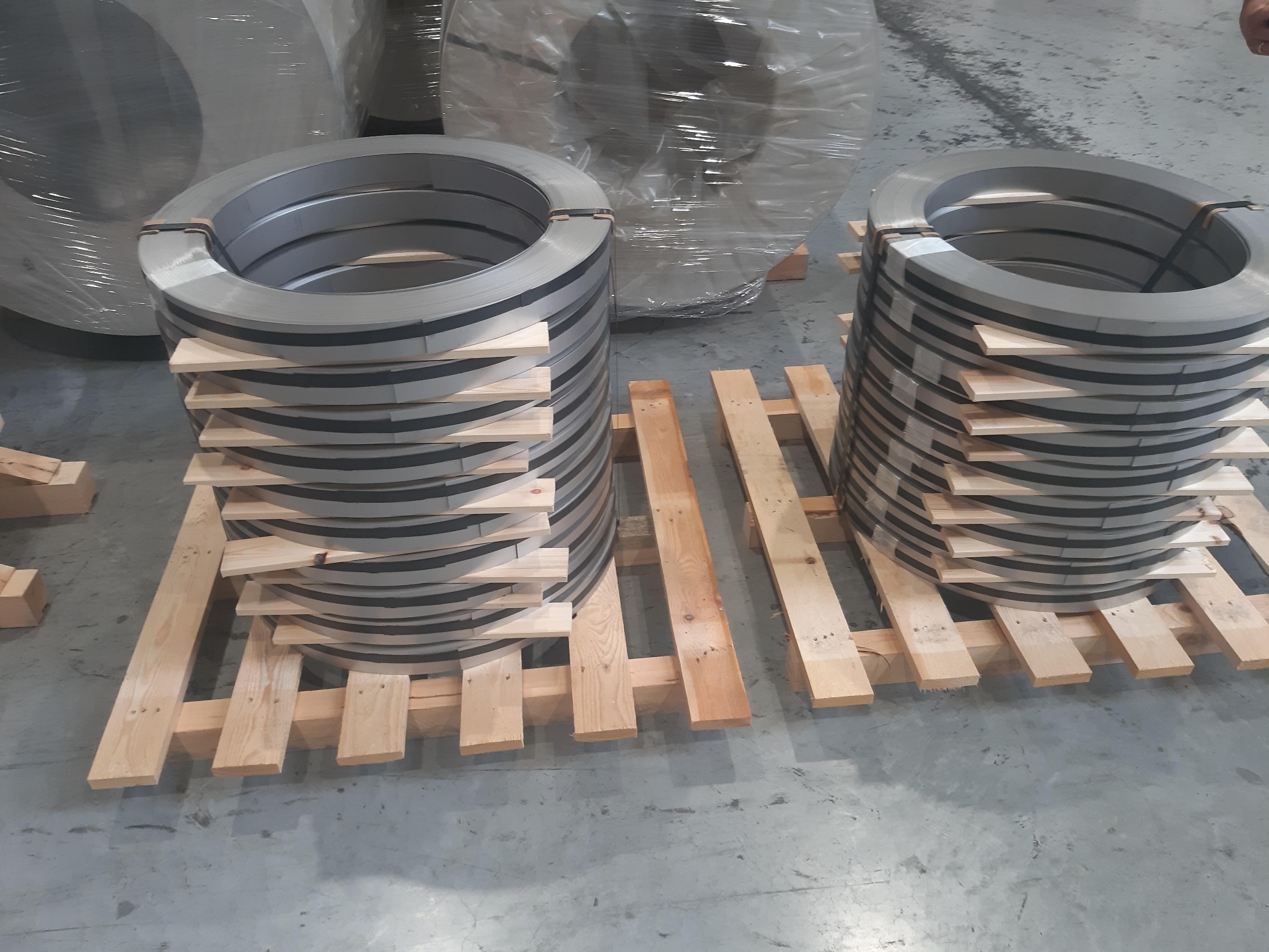 Dwie pionowe stosy zwiniętej w kręgi blachy nierdzewnej, ułożone na drewnianych paletach w magazynie.