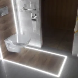 Nowoczesna toaleta podświetlona taśmą LED, z wiszącą miską WC, koszem z plecionki i fragmentem pralki w tle.