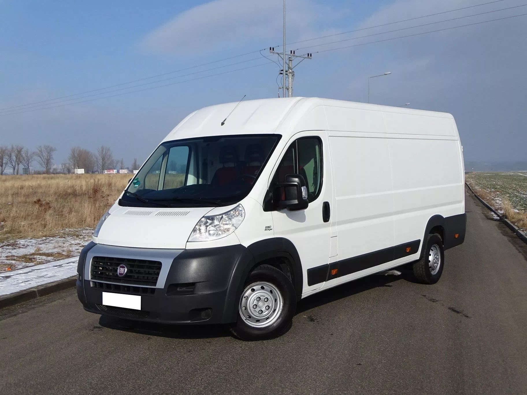 Biały samochód dostawczy Fiat Ducato zaparkowany na asfaltowej drodze, widok z przodu, w tle pole i słup energetyczny pod błękitnym niebem.