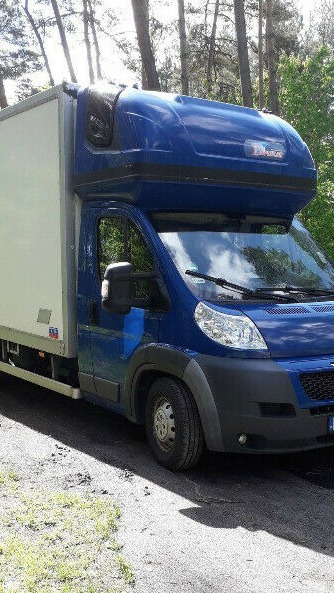 Niebieski bus transportowy z białą zabudową kontenerową, zaparkowany na poboczu drogi z widocznym lasem w tle, z charakterystycznym nadbudowanym dachem kabiny kierowcy.