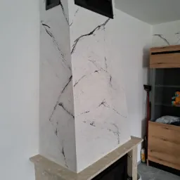 Nowoczesny kominek z obudową imitującą marmur, z czarnymi kratkami wentylacyjnymi, na tle jasnych ścian i szarej podłogi.