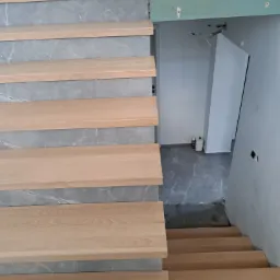 Montaż drewnianych schod&oacute;w z szarymi, marmurkowymi podstopnicami w nowym domu, buty robocze na g&oacute;rnym stopniu, widoczne przygotowania do dalszego wykończenia.